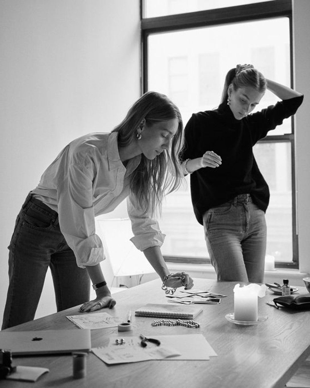 Two individuals in casual attire work at a table in a well-lit room. One leans over documents while the other stands, hand on head. A lit candle and various items are on the table.