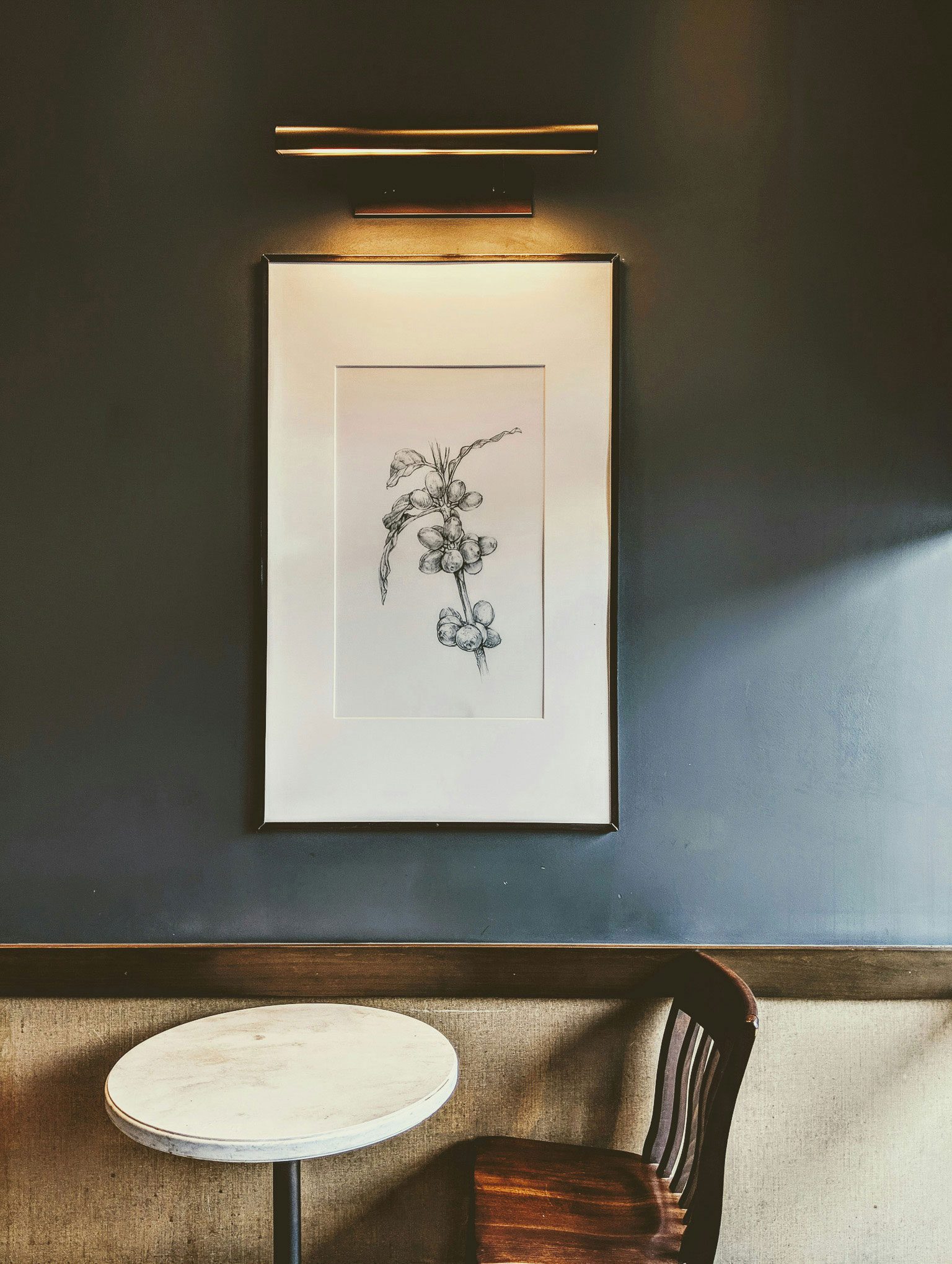 A solitary round table with a wooden chair is positioned beneath a framed botanical sketch illuminated by an overhead light on a dark-toned wall.