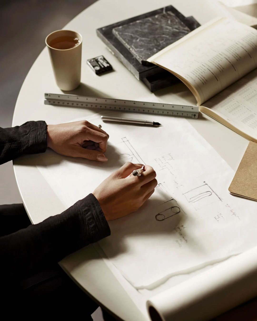 Person drawing technical sketches on a table with a ruler, book, eraser, and coffee cup.