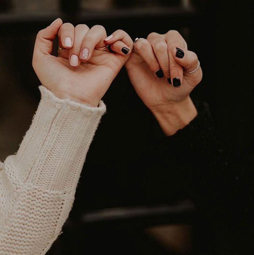 Two hands with different nail polish colors make pinky promises against a dark background. One hand has light-colored nails and a cream sweater; the other has black nail polish and a black sleeve.