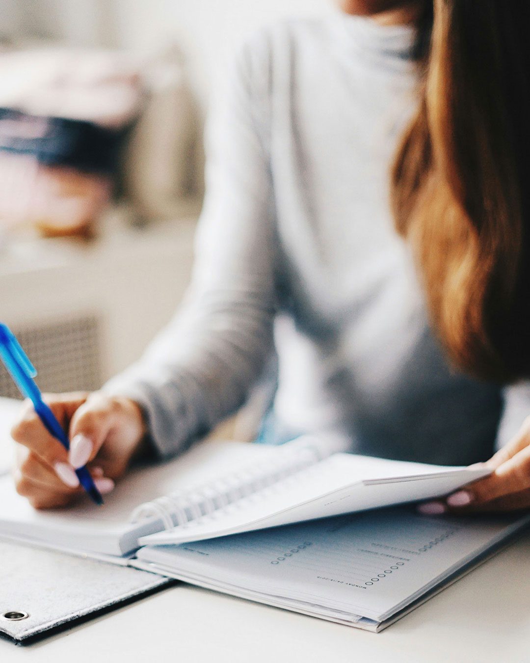 A person wearing a grey sweater writes in a notebook with a blue pen while flipping through its pages.