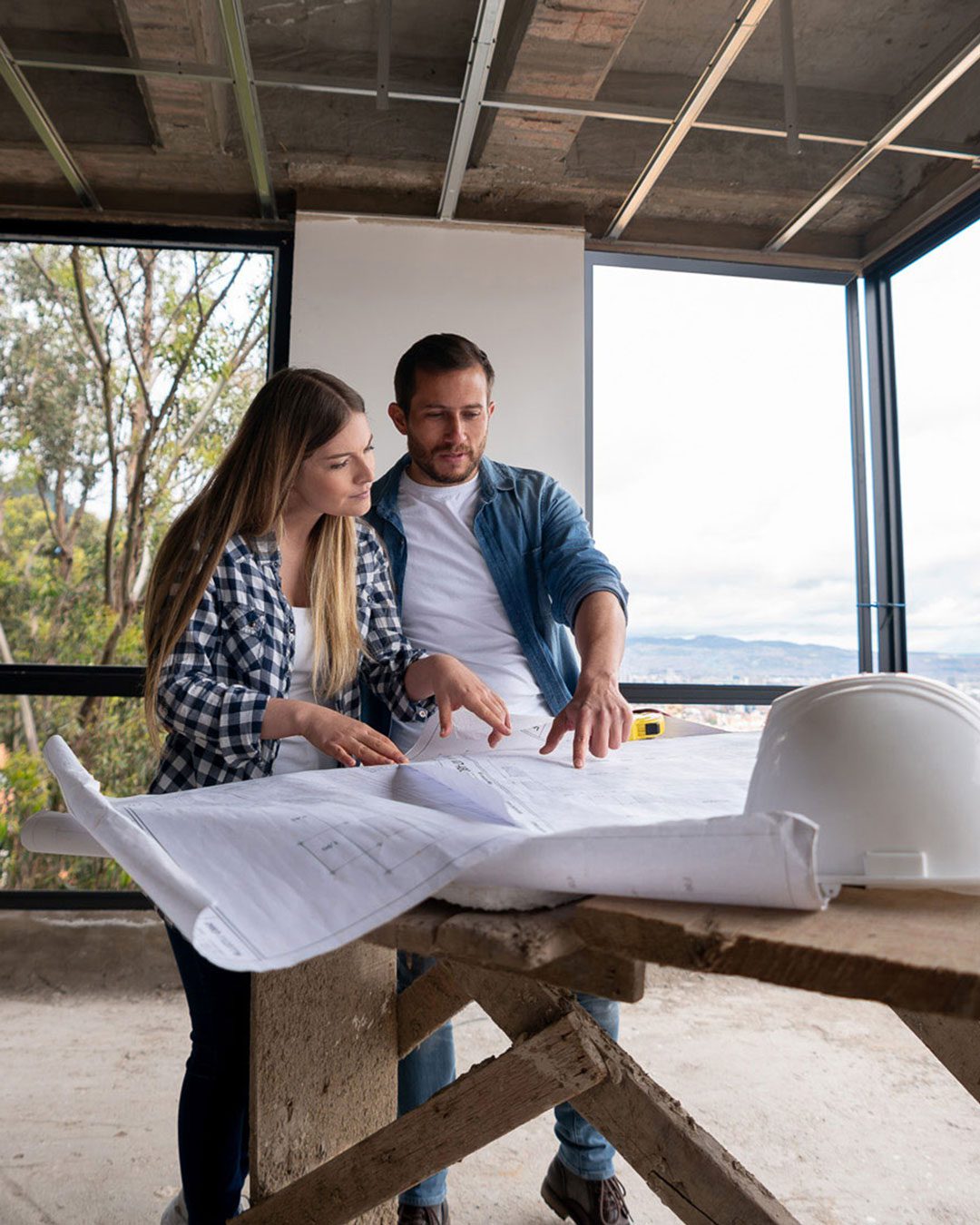 Two people standing in a partly constructed building, looking at a large blueprint on a wooden table with a white hard hat placed nearby.