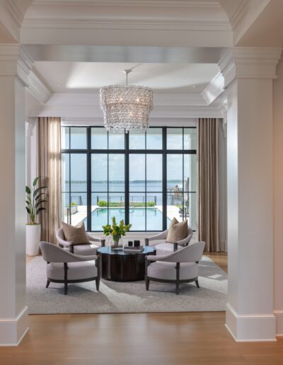 A well-lit living room with floor-to-ceiling windows, two chairs around a small table, a chandelier, and views of a swimming pool and water beyond. Walls display framed artwork.