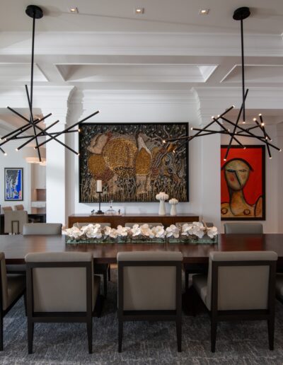 A modern dining room with a large rectangular table, ten chairs, two abstract chandeliers, and two framed artworks on white walls. The room has large windows and a coffered ceiling.