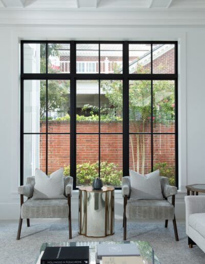 A modern living room with two grey armchairs, a small round table between them, and a large window showing a brick exterior and greenery outside. A stack of books is on a coffee table in the foreground.