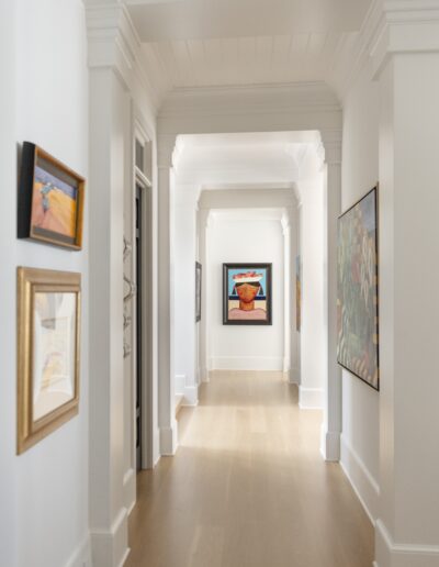 A brightly lit hallway with light wood flooring, white walls, and several colorful framed paintings hanging on both sides. The hallway leads to a room at the end with another artwork visible.