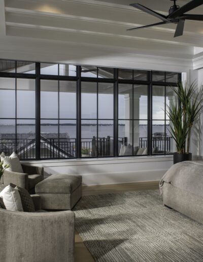 Spacious modern bedroom with large windows offering a waterfront view, featuring a king-size bed, armchairs, a potted plant, and neutral gray decor.