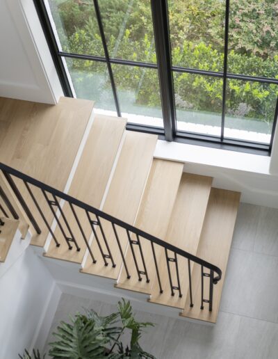 A wooden staircase with black metal railings leads to a landing by a large, grid window overlooking a garden. A potted plant sits beside the stairs.