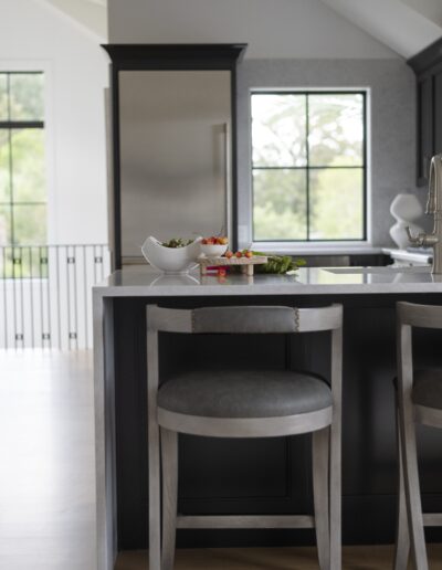 Modern kitchen with a gray island and two matching stools. Countertop features a white bowl with fruit, a small plate with vegetables, and kitchen utensils. Large windows in the background.