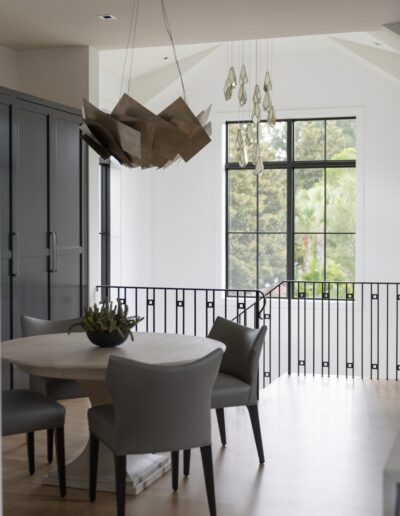 A modern dining area with a round table, four grey upholstered chairs, unique light fixtures, and large windows offering a view of greenery outside.