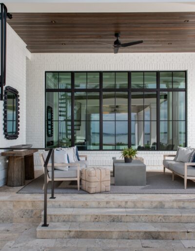 Outdoor patio with white brick walls, glass windows, a black spiral staircase, modern seating area, and wooden ceiling. Rectangular dining table with chairs in the background, overlooking a body of water.