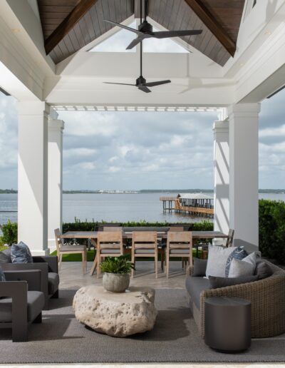 Covered outdoor patio with seating area, wooden dining table, and ceiling fans overlooking a large expanse of water and a distant pier.